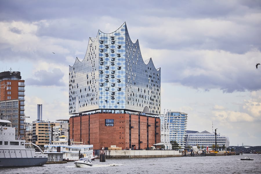 Elbphilharmonie in Hamburg, filigraner Glasbau auf einem massigen, rötlichen Kaispeicher an der Elbe, Ansicht vom Wasser aus, mehrere Schiffe links von der Elbphilharmonie, andere Gebäuden der HafenCity neben der Elbphilharmonie links und hinter der Elbphilharmonie rechts