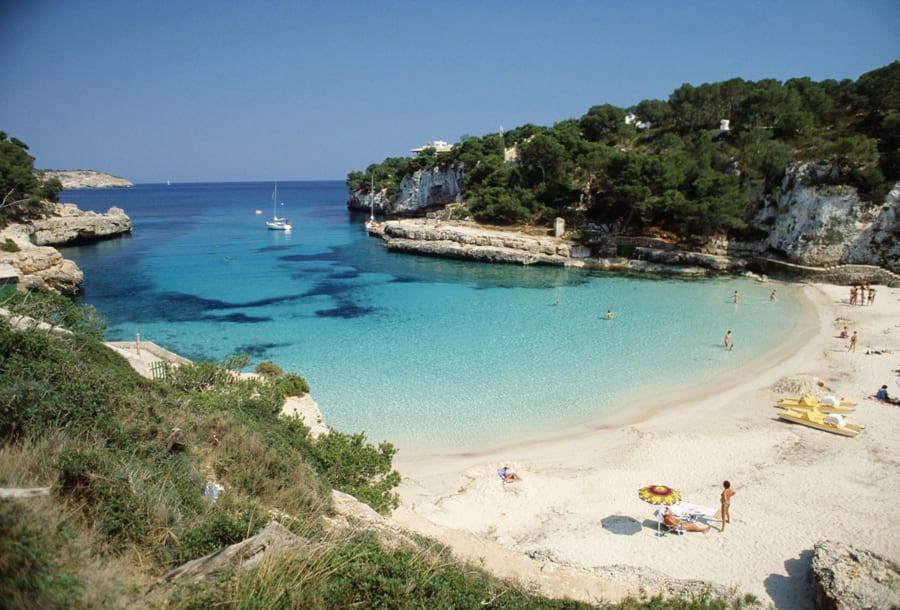 Mallorca. Blick von oben auf die traumhafte Bucht Cala Llombards mit feinem Sandstrand, türkisblauem Meer umrahmt von zerklüfteten, begrünten Felsformationen. Blauer Himmel, Sonne