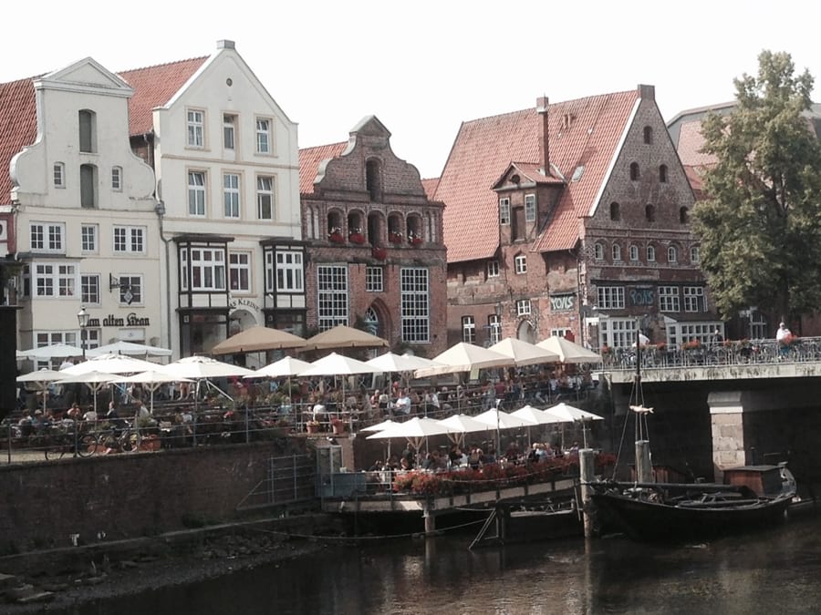 Idyllisches Lüneburg am alten Hafen, Restaurants Außenbereiche mit Sonnenschirmen im Vordergrund, schmucke Patrizierhäuser mit roten Backsteinfassaden und weißen Fassaden dahinter. Ein alter Segel-Frachtkahn liegt im Hafen.
