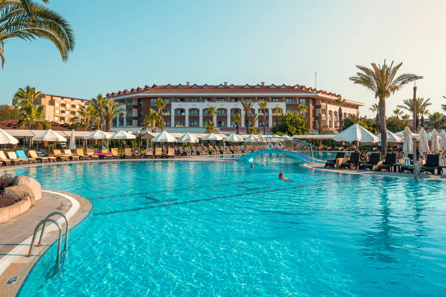 Club Hotel Turan Prince World Select Villa mit großem Swimmingpool davor mit Liegen, Sonnenschirmen und Palmen. Sonne, blauer Himmel