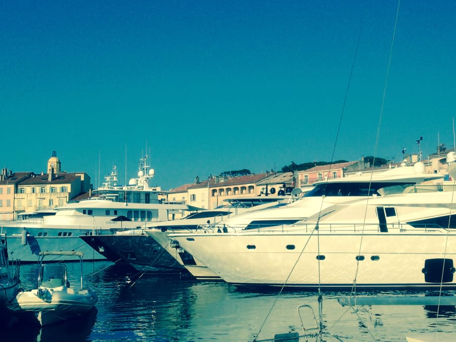 Große Luxus-Yachten im Hafen von Saint-Tropez liegen dicht nebeneinander, dahinter die typischen orangefarbenen und gelben provenzalischen Häuser von Saint-Tropez. Blauer Himmel, Sonne
