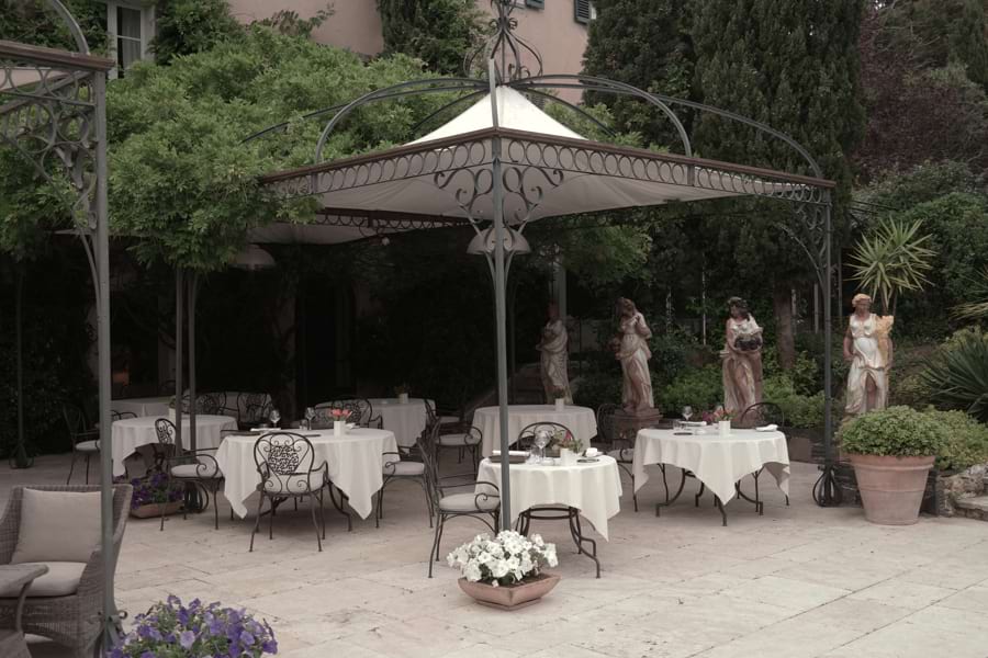 Restaurant La Table du Mas auf der Terrasse draußen, mehrere runde Tische schön eingedeckt mit edlem Geschirr, Weingläsern und weißen Tischdecken unterm Baldachin. Göttinnen-Statuen im Hintergrund, grüne Bäume und Büsche im Hintergrund