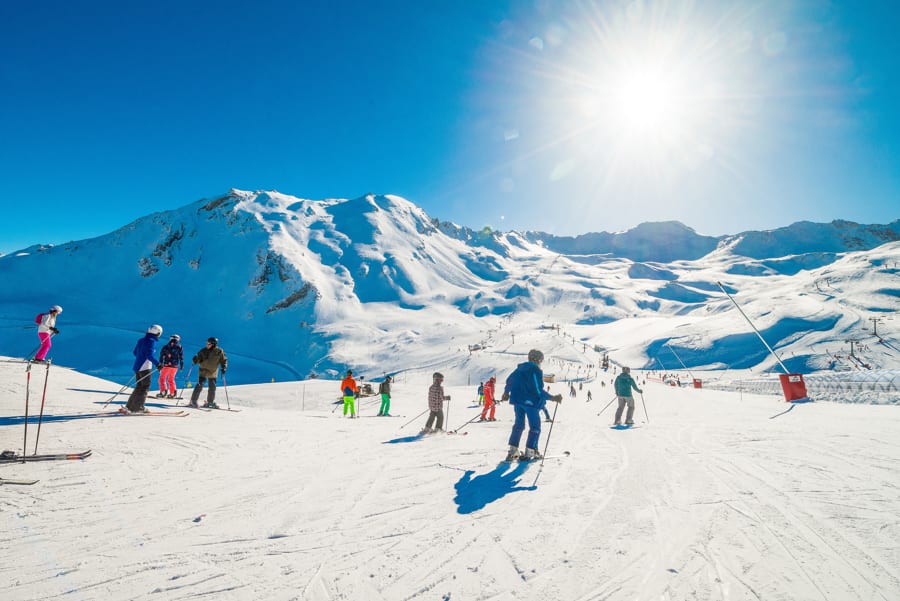 Auf schöner, breiter Skipiste fahren einige Skifahrer auf gutem Schnee runter mit Blick auf das herrliche, schneebedeckte, weite Gebirgspanorama mit Skiliften im Hintergrund mit strahlender Sonne und blauem Himmel.