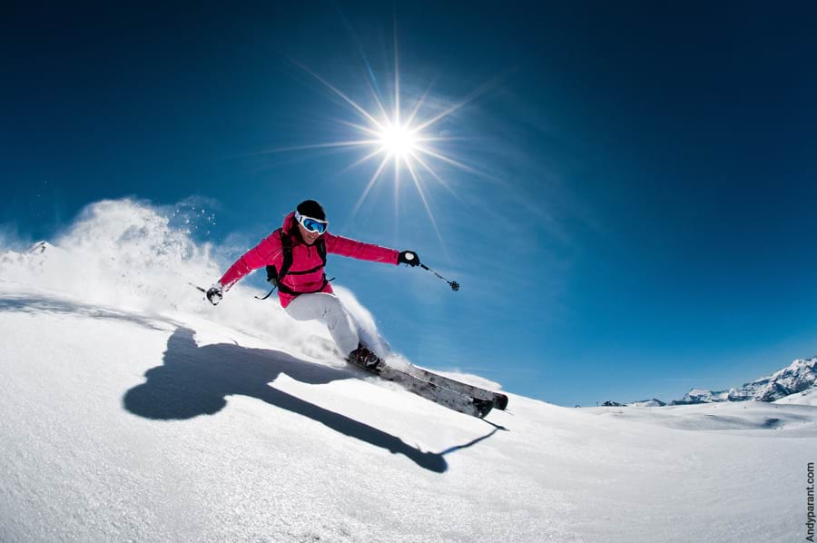 Guter Skifahrer fährt rasant mit perfekter Skitechnik auf einer steilen, anspruchsvollen Piste mit gutem Schnee. Schnee staubt hinter ihm auf. Die Sonne scheint strahlend am blauen Himmel.