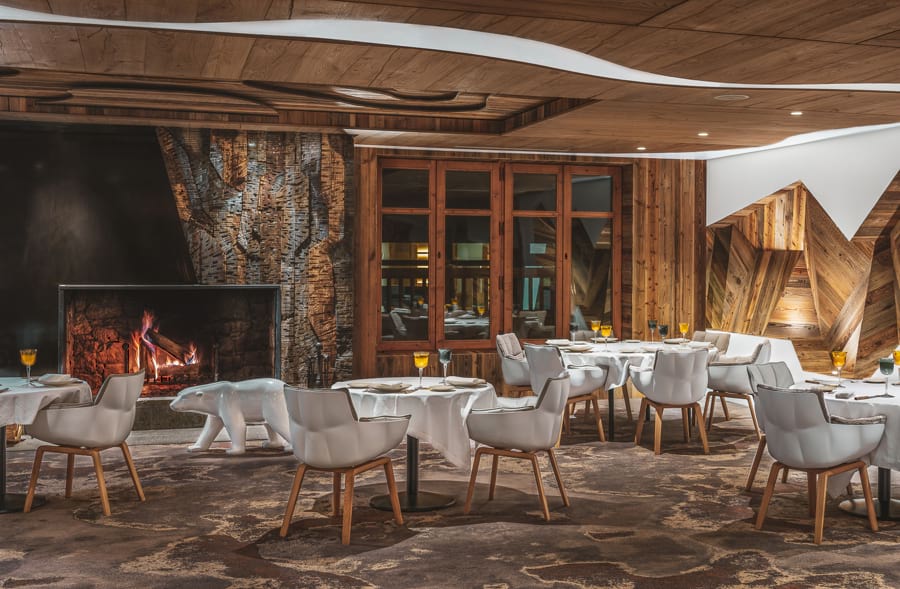 Blick ins Restaurant La Table de l'Ours: exklusives, verführerisches Ambiente: schönes, braunes Holz an den Wänden, imposante Holz-Deckenverkleidung in Braun, großer, lodernder, knisternder Kamin, weiße Stühle an weiß eingedeckten Tischen