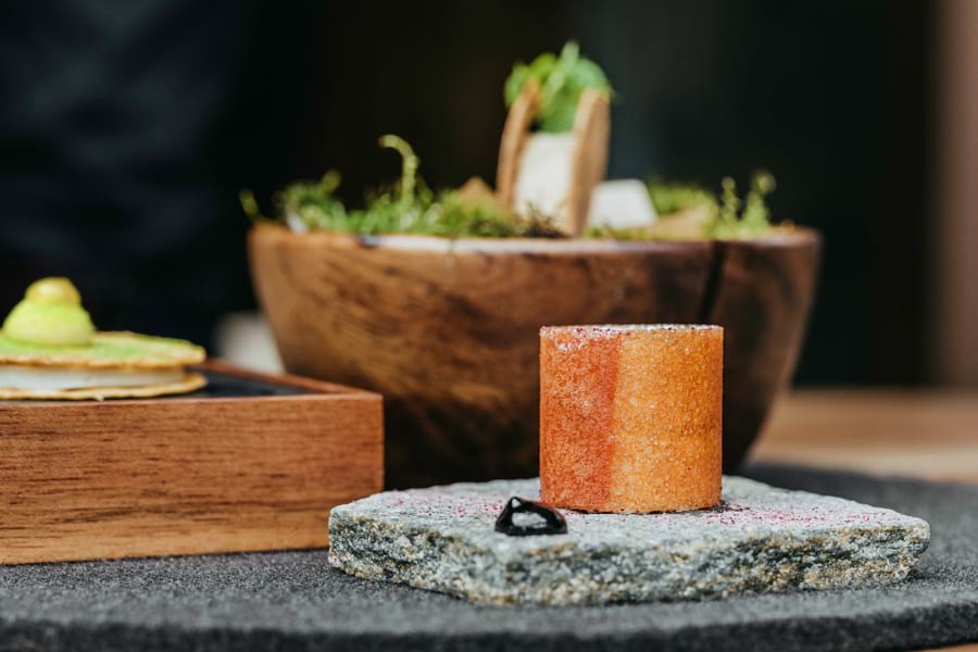 Restaurant L’Atelier d’Edmond: Gericht in Zylinder-Form in Gelb-Orange steht auf grauer Granitsteinplatte, dahinter braune Holzschale mit Gemüse
