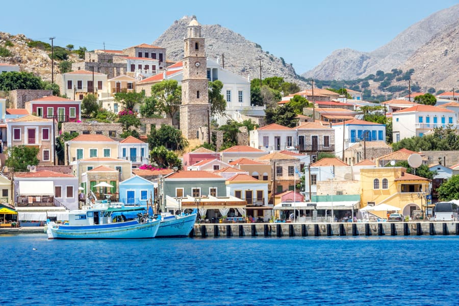 Insel Rhodos in Griechenland Blick vom blauen Meer im Vordergrund auf griechische Häuser mit gelben, orangefarbenen, blauen Fassaden am Hafen mit zwei blauen Fischerbooten am Kai davor. Die Sonne scheint, blauer Himmel.