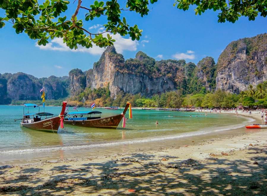 Feinsandiger Strand, grünblautürkisfarbenes Meer, idyllische Bucht, zwei thailändische Ausflugsboote aus braunem Holz ankern in der Bucht, Sonne, blauer Himmel, grüne Bäume am Strand, hohe graue Felsen im Hintergrund