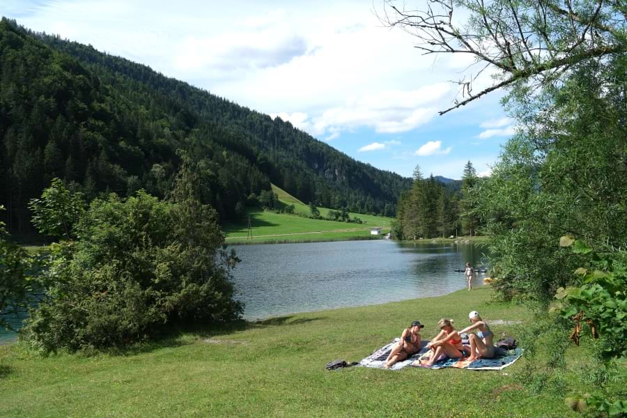 Smaragdgrüner Pillersee eingerahmt in idyllischer Berglandschaft mit grünen Nadelbäumen und großer Liegewiese. 3 junge Frauen sonnen sich auf ihren Badetüchern auf der Liegewiese