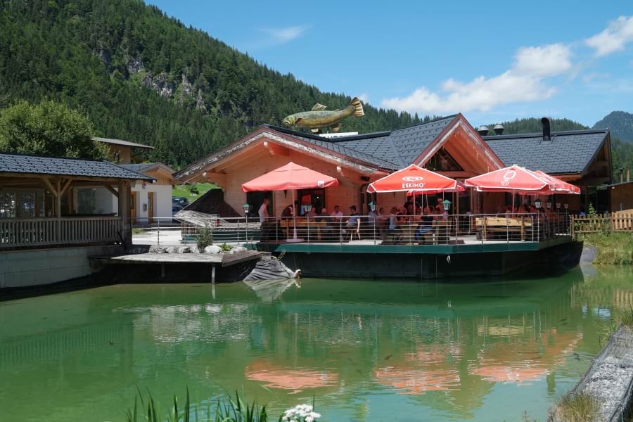 Forellenranch Restaurant am Forellenteich, schöne Terrasse mit roten Sonnenschirmen, eine große Forelle ist als Werbesymbol auf dem Dach zu sehen