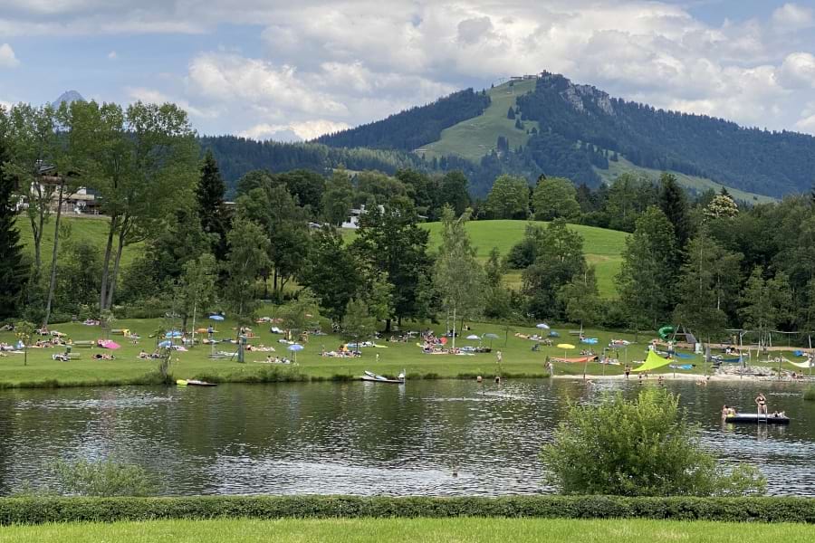 Lauchsee mit Badenden. Auf der grünen Liegewiese liegen Badegäste. Grüne Bäume hinter der Liegewiese, grüne Wiesen, Berge mit grünen Bäumen und grünen Hängen