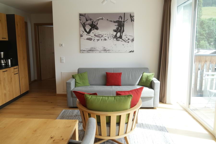 Schickes, geradliniges, bequemes Sofa mit grauem Stoff und breiter Sessel aus hellbraunem Holz mit roten und grünen Kissen im Luxus-Apartment im All-Suite Resort Fieberbrunn