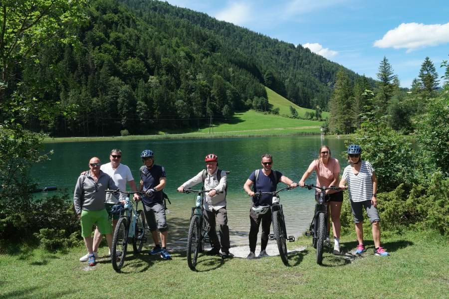 E-Bike-Fahrer der Pressereise-Gruppe stehen mit ihren E-Bikes vor dem smaragdgrünen Pillersee in einer idyllischen Berglandschaft mit grünen Nadelbäumen