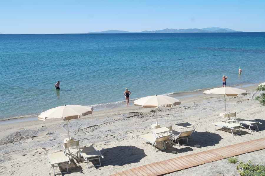 Blick von oben auf den Privatstrand vom The Sense Experience Resort Hotel. Drei Sonnenschirme mit jeweils zwei Liegen und Regiestuhl alles in der Farbe Hellbeige auf feinem, hellen Sand vorn im Bild. Dahinter liegt das blaue, weite Meer. Die Sonne scheint, hellblauer Himmel.
