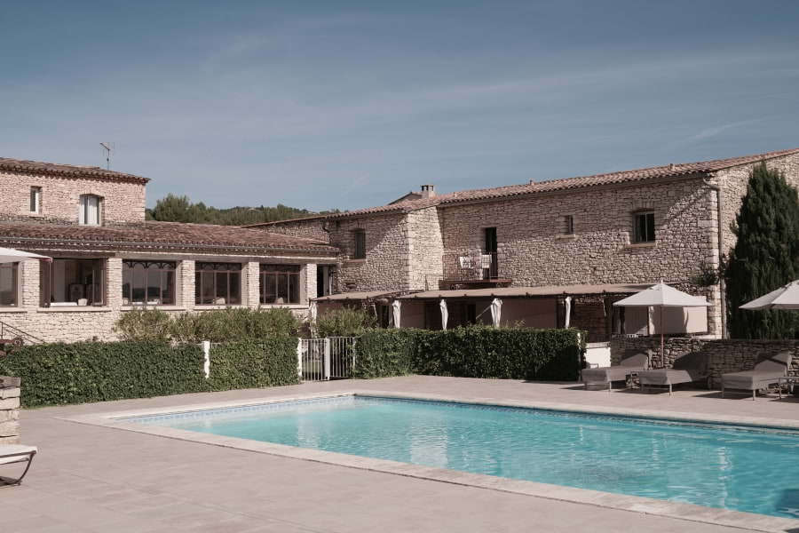 Hotelanlage Le Phébus & Spa in roher Architektur der Provence aus hellen Trockensteinen. Davor grüne Hecke, davor Swimming Pool mit grauen Liegen und weißen Sonnenschirmen. Blauer Himmel, die Sonne scheint.