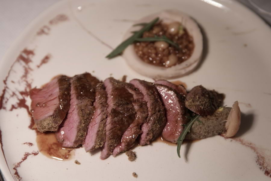 Aufgeschnittene, rosa Lammkeule mit brauner Soße liegt auf großem, weißen Teller. Sieht lecker aus. Gemüse-Frikassee aus Einkorn und Erbsen mit brauner Soße liegt in einer Schale hinter der Lammkeule auf dem Teller.