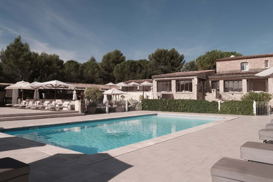Hotelanlage Le Phébus & Spa in roher Architektur der Provence aus hellen Trockensteinen mit dem Michelin-Stern-Restaurant La Table de Xavier Mathieu mit großen Fenstern. Davor grüne Hecke, davor großer Swimming Pool mit weißen Liegen und weißen Sonnenschirmen. Direkt hinter dem Hotel beginnt der üppige Wald mit vielen grünen Bäumen. Blauer Himmel, die Sonne scheint.