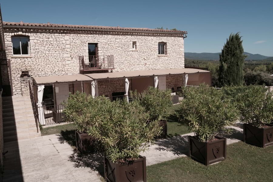 Faszinierendes Le Phébus & Spa Luxushotel im provenzalischem Baustil mit hellen Trockensteinen. Eigene Terrassen mit Sonnenschutz. Davor stehen viele grüne Büsche auf grünem Rasen. Blauer Himmel, die Sonne scheint.