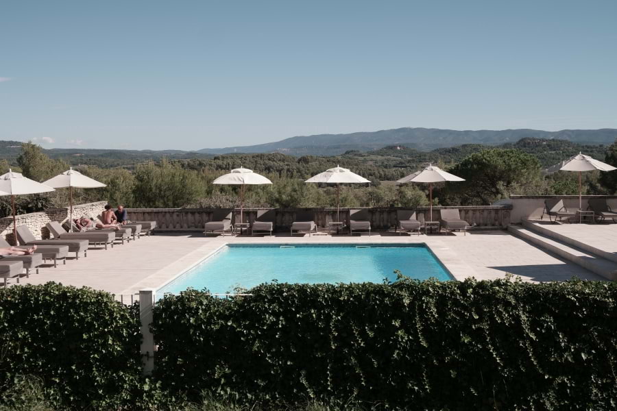 Schöner großer Swimming Pool mit schicken, grauen Liegen und weißen Sonnenschirmen. Wunderschöner Blick ins Tal auf die idyllische, grüne Landschaft vom Luberon. Vor dem Swimming Pool steht eine grüne, niedrige Hecke. Blauer Himmel, die Sonne scheint.