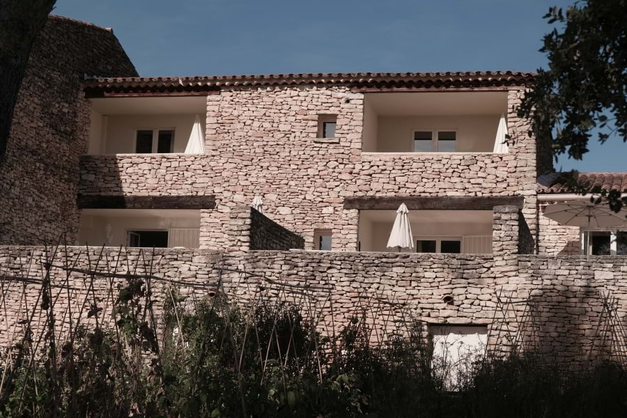 Hotel Le Phébus & Spa in roher, charakteristischer Architektur aus hellen Trockensteinen im Baustil der Provence. Außenansicht auf vier Zimmer mit großen Terrassen oder Balkonen. Blauer Himmel, die Sonne scheint.