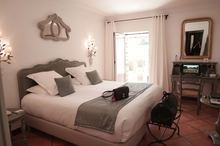 Provenzalisch luxuriöses Zimmer im Le Phébus & Spa Luxushotel mit sanften Farben wie Weiß, Grau und natürlichen Materialien wie Holz, Stein, Schmiedeeisen eingerichtet. Großes, komfortables Doppelbett mit weißer Bettdecke, weißen Kissen und hoher Matratze. Rustikaler Schreibtisch aus grauem Holz steht rechts an der Wand. Rötlichbraune große Bodenfliesen. Durch ein großes Fenster fällt Licht ins Zimmer. Weiße Vorhänge hängen neben dem Fenster
